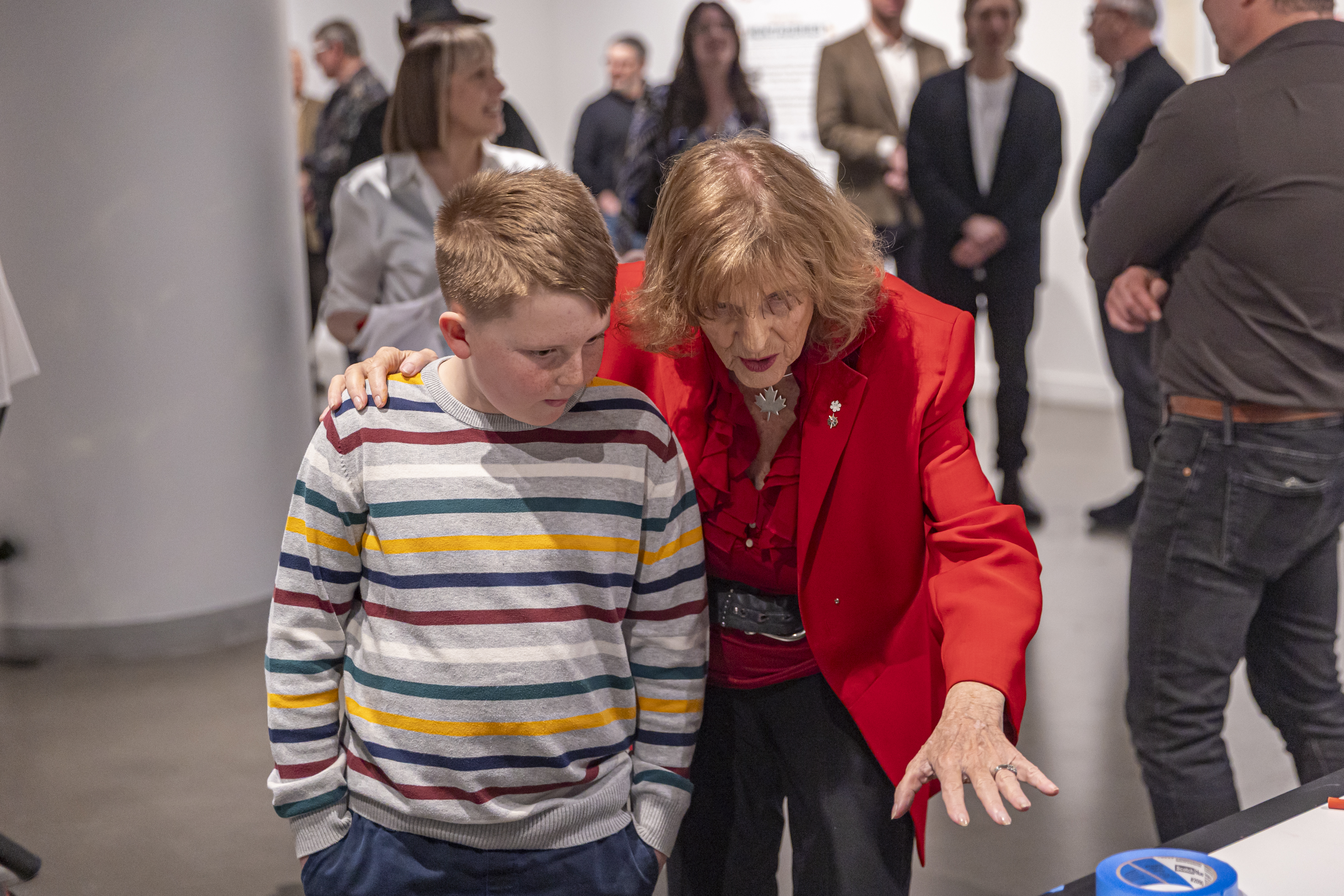 Former Lieutenant Governor of Alberta, wearing a red blazer, interacts with a young boy wearing a yellow, blue, green, red, and white striped sweatshirt.