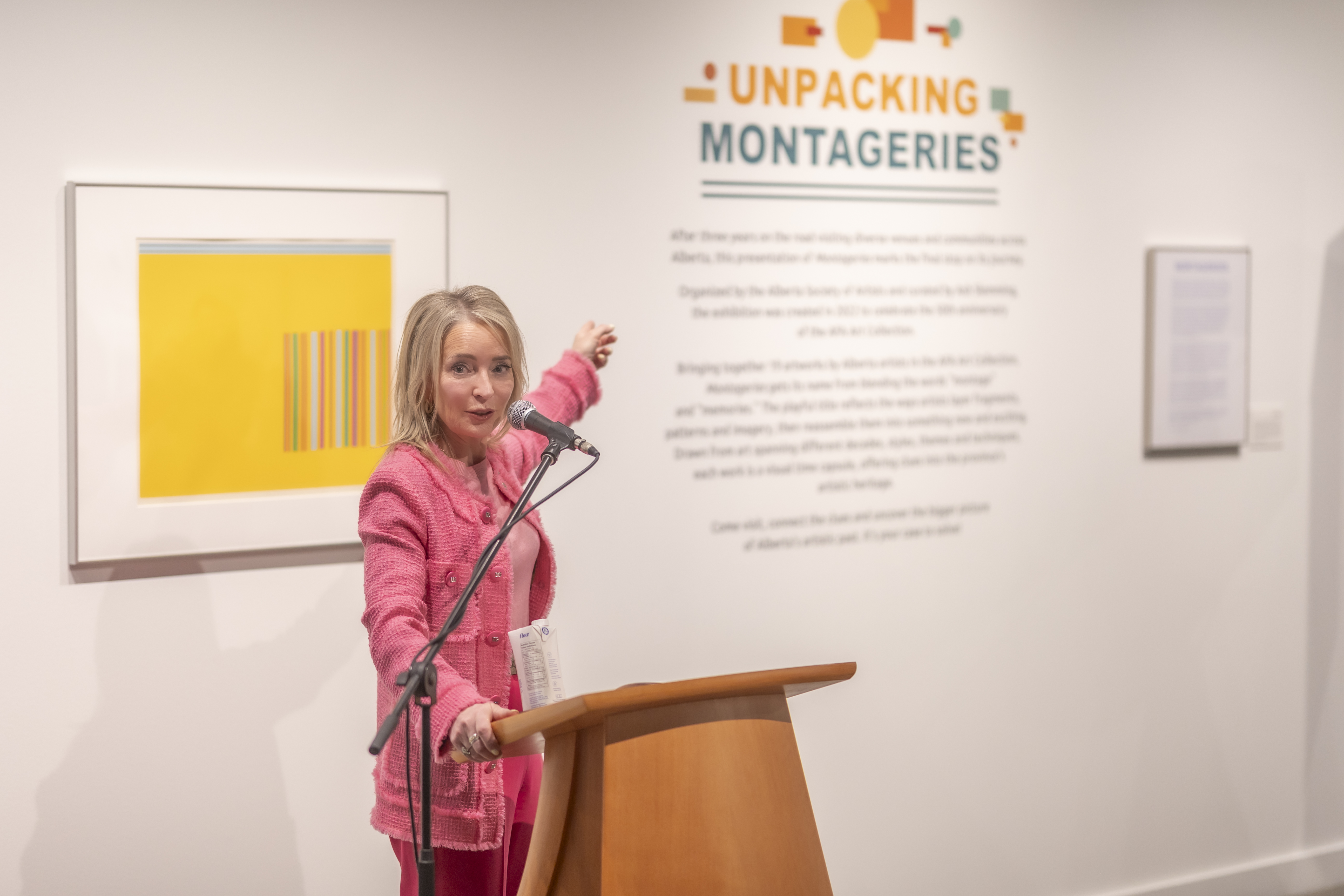 Cynthia P. Moore speaks behind a brown podium and gesgtures towards the Unpacking Montageries display on the wall. Moore is wearing a pink blazer