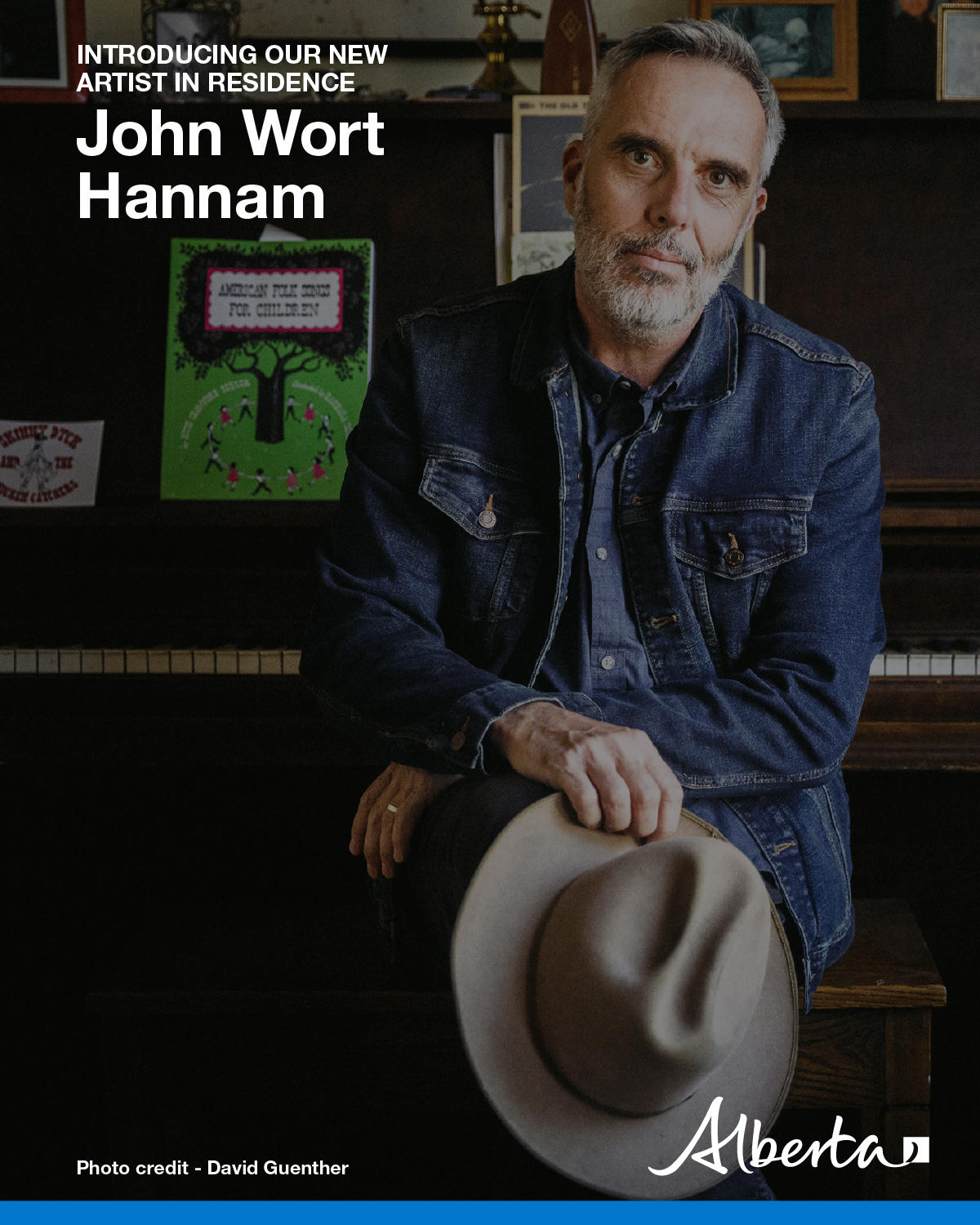 Man with short white beard in jean jacket an holding white cowboy hat leans against an upright piano