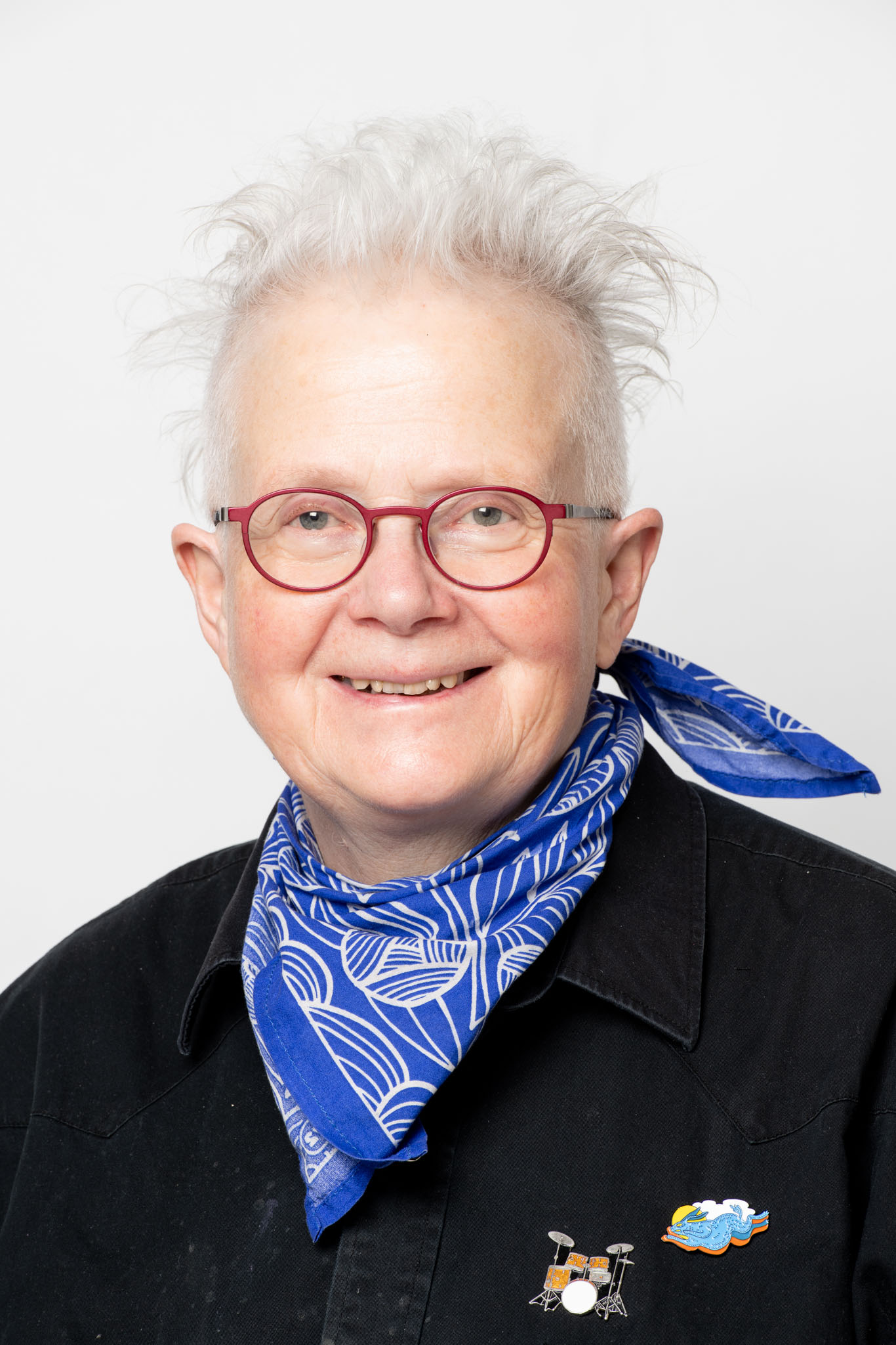 Portrait of artist Rita McKeough. She has short white hair, glasses, and is wearing a blue scarf and dark jean jacket.