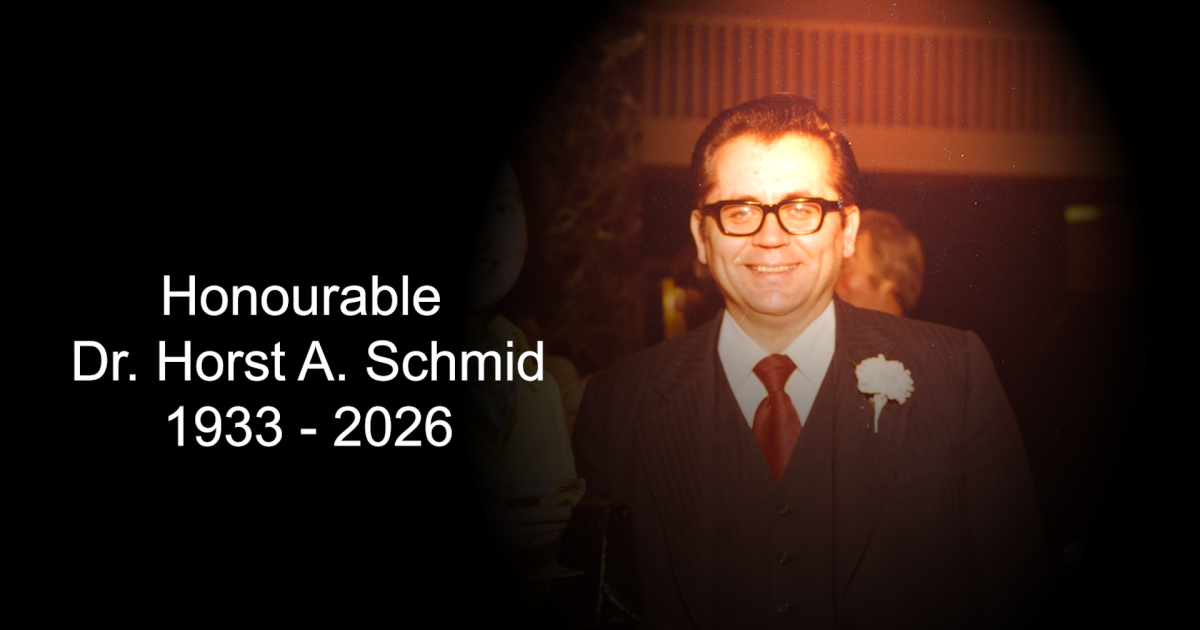 An archival photo of Horst Schmid, who is wearing glasses with a suit, red tie, and a corsage pinned to his suit jacket. Text accompanying the photo reads Honourable Dr. Horst A. Schmid 1933 – 2026.