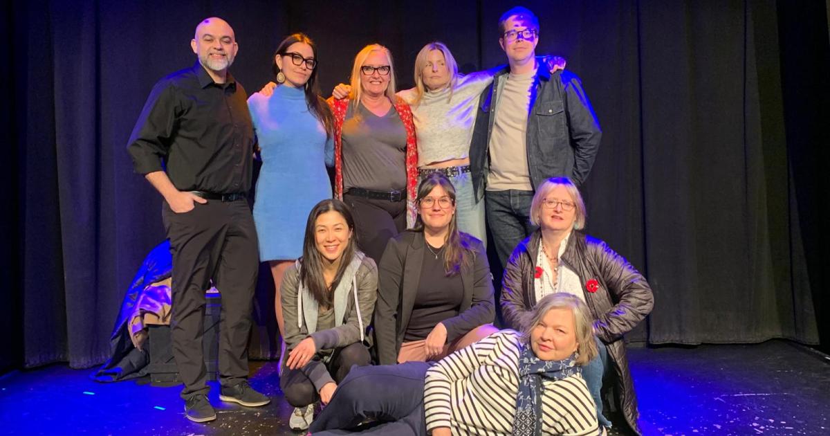 Nine comedians in a theatre space with black curtains pose and smile.