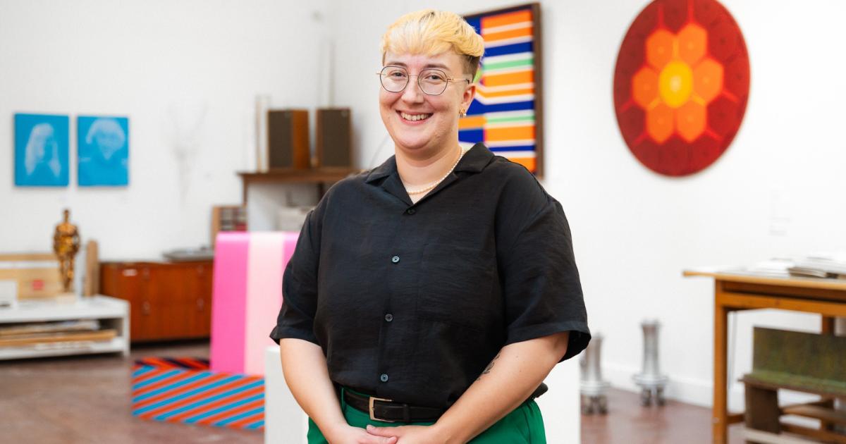 Ash Slemming is photographed standing and smiling while wearing a black-collared shirt and green pants. A number of artworks can be seen hanging on the wall in the background. Photo credit: Austin Kamenz.