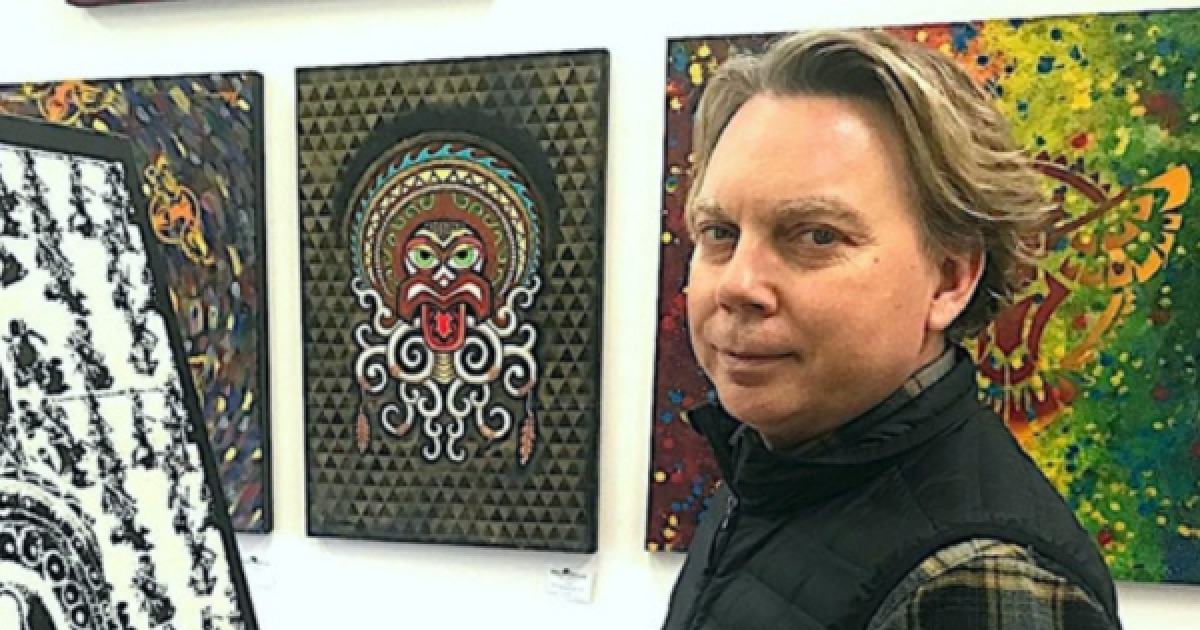 AFA board member Jason O’Connor stands in front of an artwork facing the camera while wearing a black vest with a plaid shirt underneath. Three other artworks line the wall behind him.