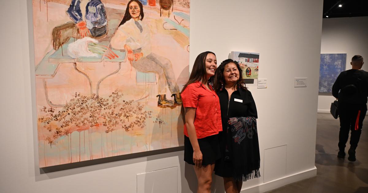 Kiona Callihoo Ligtvoet and Maureen Callihoo pose side by side beside their respective artworks. Kiona is wearing a red blouse and black skirt while Maureen is wearing a black jacket.